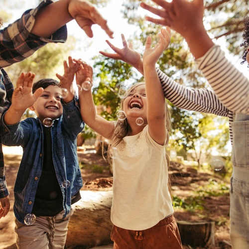Four children laugh and smile as they play outside together. The group is trying to catch bubbles in their hands.
