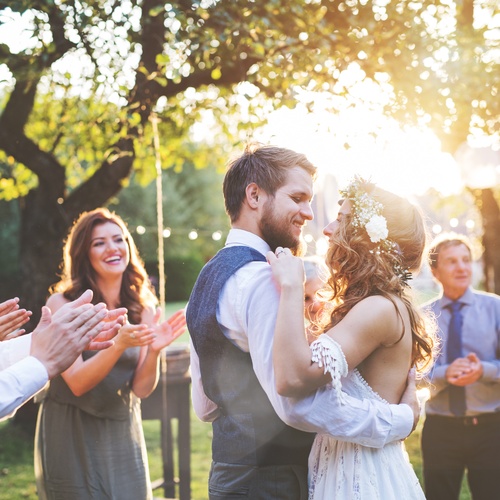 A bride and groom dancing at their outdoor wedding ceremony. The sun shines on them, and guests cheer them on.