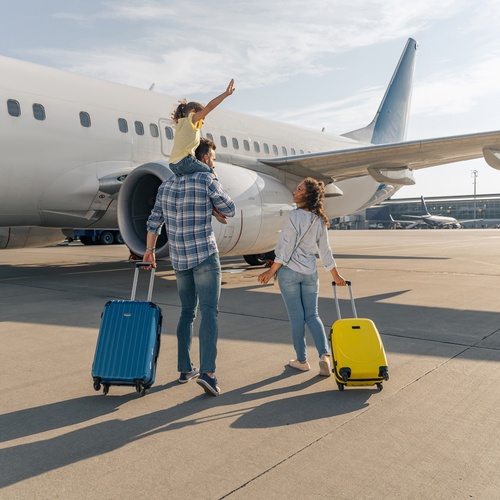 A man and woman with suitcases walk toward an airplane outside. The man carries a child on his shoulders.