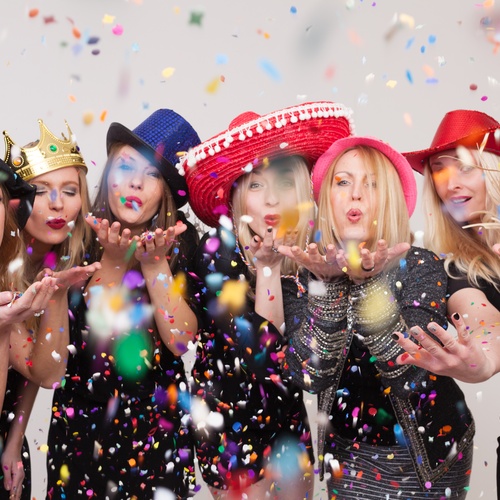 Close-up of seven woman posing for a photo while wearing hats and silly costumes and throwing handfuls of confetti.
