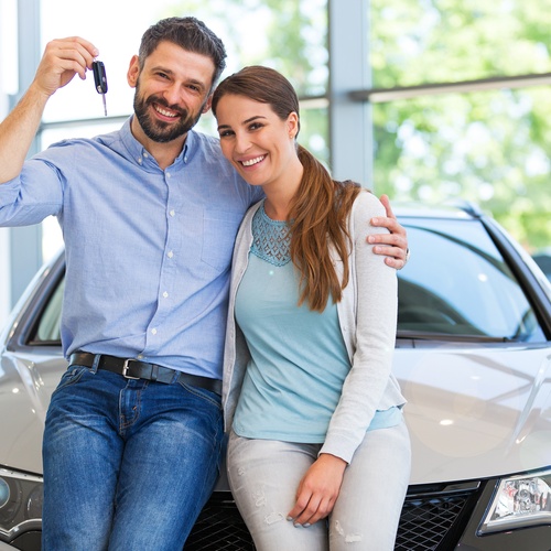 A young couple stands and smiles at a car dealership, holding the new car keys because they bought a new car.