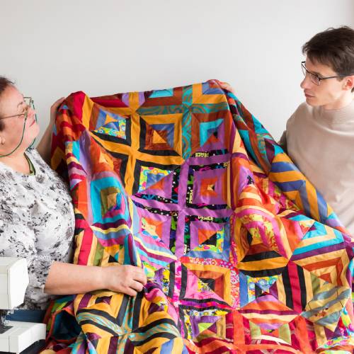A man and a woman stand next to a sewing machine, holding up a colorful homemade quilt that they made.