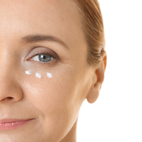 A close-up of the left side of a middle-aged woman's face. She has three dots of moisturizer under her eye.
