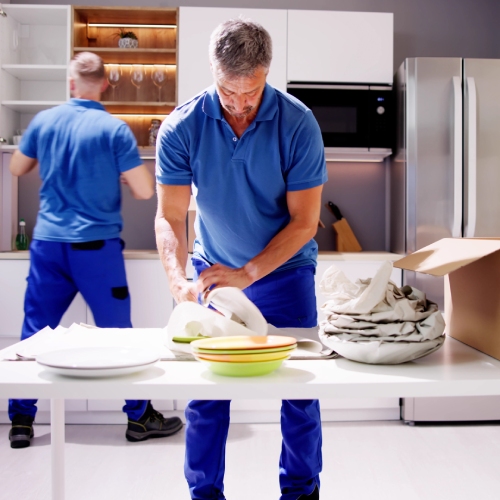 A packer wraps dishes with paper before placing them in a cardboard box. Another worker empties a cabinet in the background.