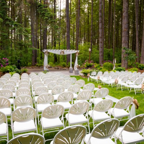 An outdoor wedding ceremony is set up in the forest with many white chairs and a white square arch at the front.