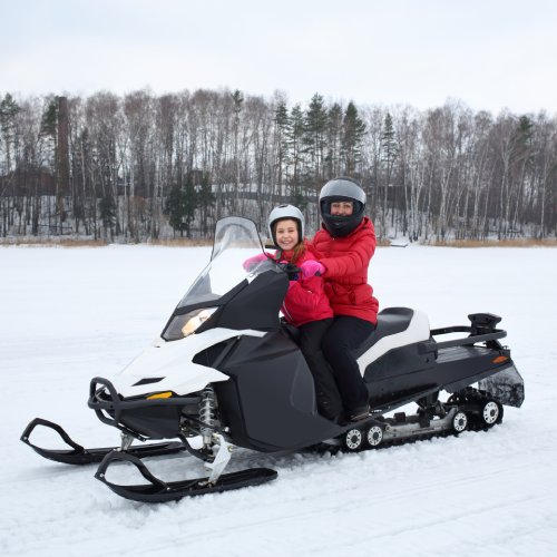 A mother and her young daughter sit on a snowmobile in a field. Both wear helmets and bright red winter coats.