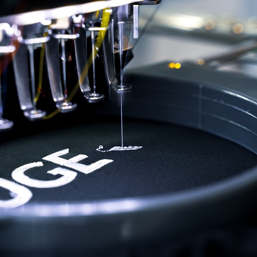 A close-up of an embroidery machine embroidering words in white letting onto a piece of black fabric.
