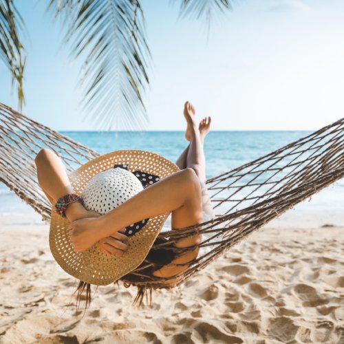 A person wearing a sun hat lounging in a hammock over the sand on the beach, near a palm tree and facing the ocean.