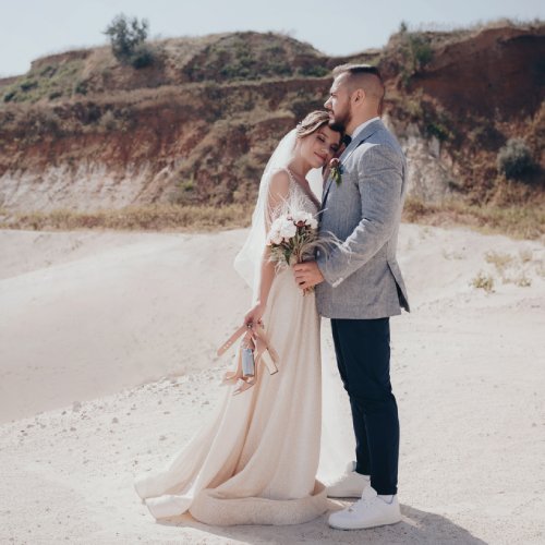 Happy newlyweds face one another while standing on a beach. The bride rests her cheek on the groom's chest.