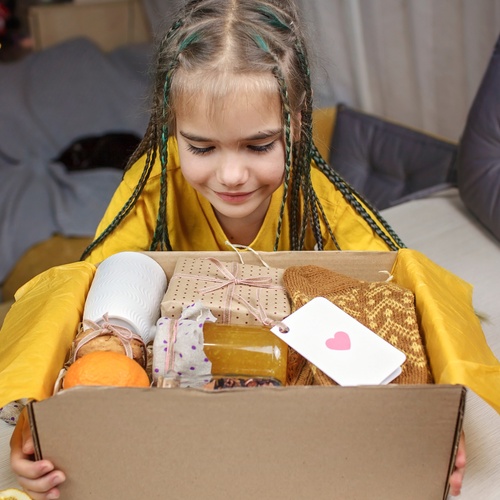 A little girl in a yellow shirt holding an open cardboard box full of socks, food, and other wrapped gifts.