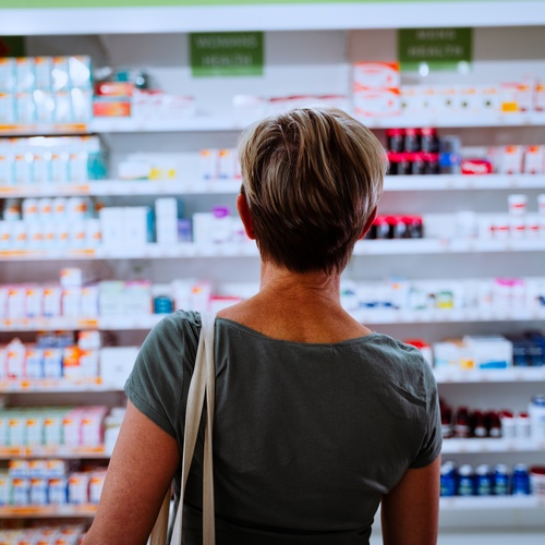 A woman wearing a green T-shirt faces a wall of shelving full of products in a drugstore pharmacy.