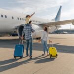 A man and woman with suitcases walk toward an airplane outside. The man carries a child on his shoulders.
