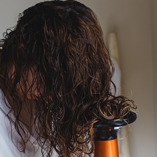 A close-up of a woman using a diffuser to blow dry her curly hair. She's wearing a lightweight light colored shirt.