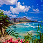 Waikiki beach with the Honolulu skyline and Diamond Head volcano in the distance on a partially cloudy sky.