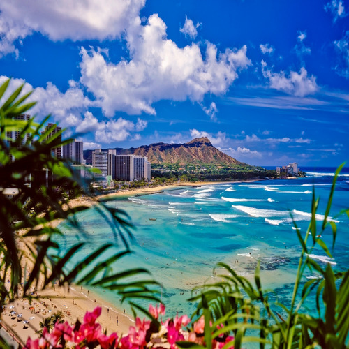 Waikiki beach with the Honolulu skyline and Diamond Head volcano in the distance on a partially cloudy sky.