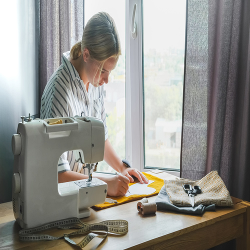 A woman is using a sewing machine and an older textile to create a fabric good. She's using a stencil on the yellow fabric.
