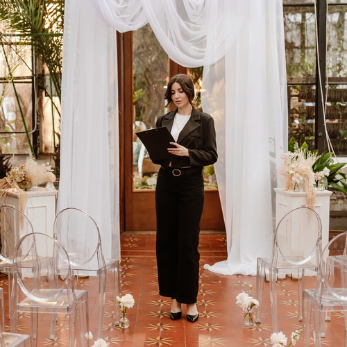 A woman wearing a black skirt and jacket writes on a clipboard. She stands in front of rows of clear chairs and draped fabric.
