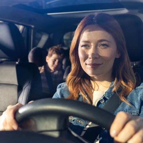 A woman driving her car with a smirk on her face as she drives toward the sun. There is a child in the backseat.