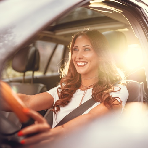 A woman driving her new vehicle with a smile on her face. The sunlight is shining behind her as she looks forward.