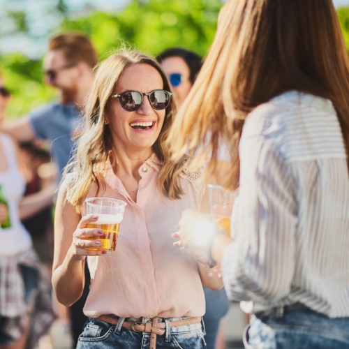 woman in sunglasses holds a clear cup full of a drink and talks to another woman outside in front of other people.