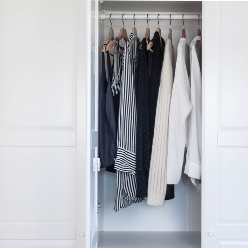 An open, white closet door, revealing navy, black, ivory, and white tops, including tank tops and long-sleeve shirts.