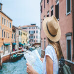 A woman in a short-sleeve shirt and a sun hat near a canal, holding a paper map with a camera strapped to her back.