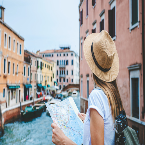 A woman in a short-sleeve shirt and a sun hat near a canal, holding a paper map with a camera strapped to her back.