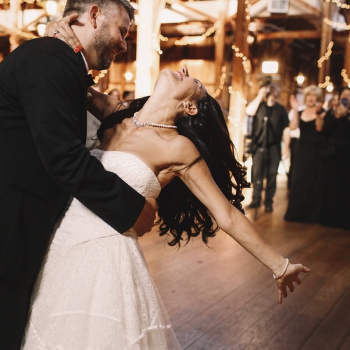 A groom in a tux dips his bride while dancing. She's wearing a strapless dress and her brunette hair is loose around her.