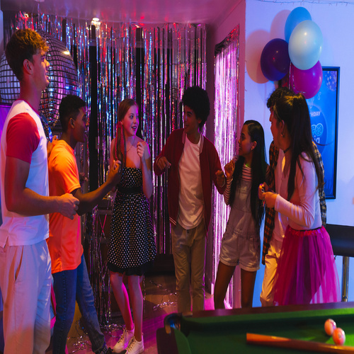 A group of diverse teen friends dancing and laughing at a vibrant party with decorations and balloons at home.