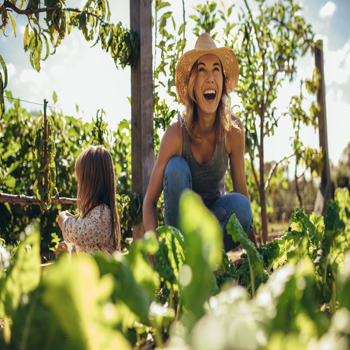 A mother and daughter are outside gardening together. The mother is smiling while planting romaine lettuce.