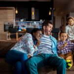 Man and woman sitting next to a young boy on a couch while two girls run to them inside the living room of a house.
