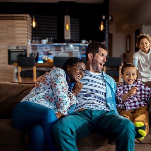 Man and woman sitting next to a young boy on a couch while two girls run to them inside the living room of a house.