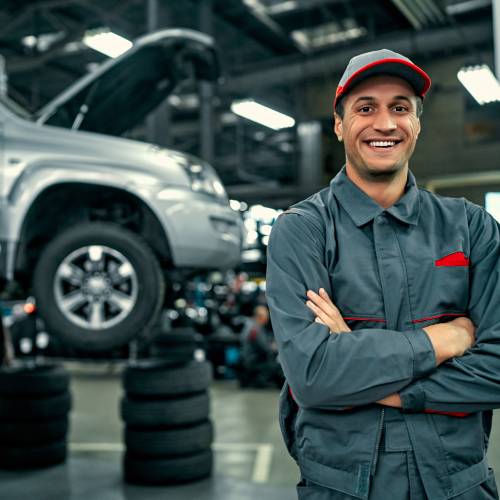 An auto mechanic in uniform stands next to a car with its hood open, smiling and looking straight into the camera.