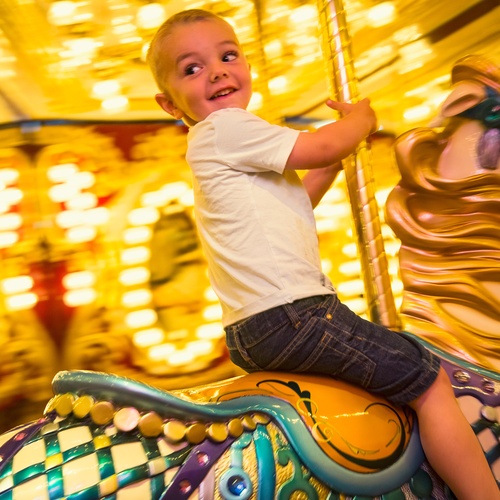 A young boy sits on top of a decorative horse on a carousel. There are bright yellow lights and mirrors in the background.