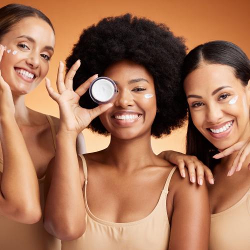 Three diverse women are posing with beauty products and smiling in front of a brownish-orange background.