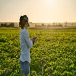 A woman farmer checks the field with a tablet. She's standing in the middle of green crops next to a sprinkler.