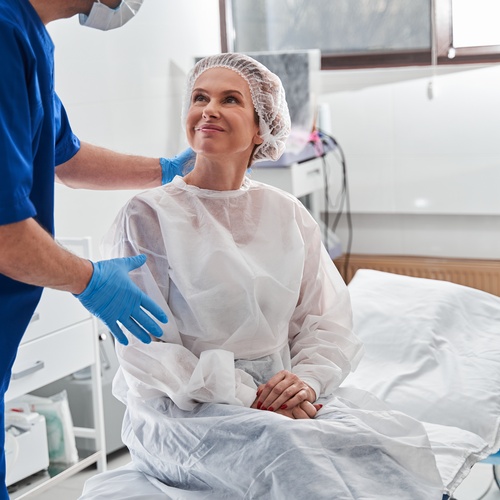 A mature woman is dressed and prepared for surgery. She's smiling and talking to the male medical professional.